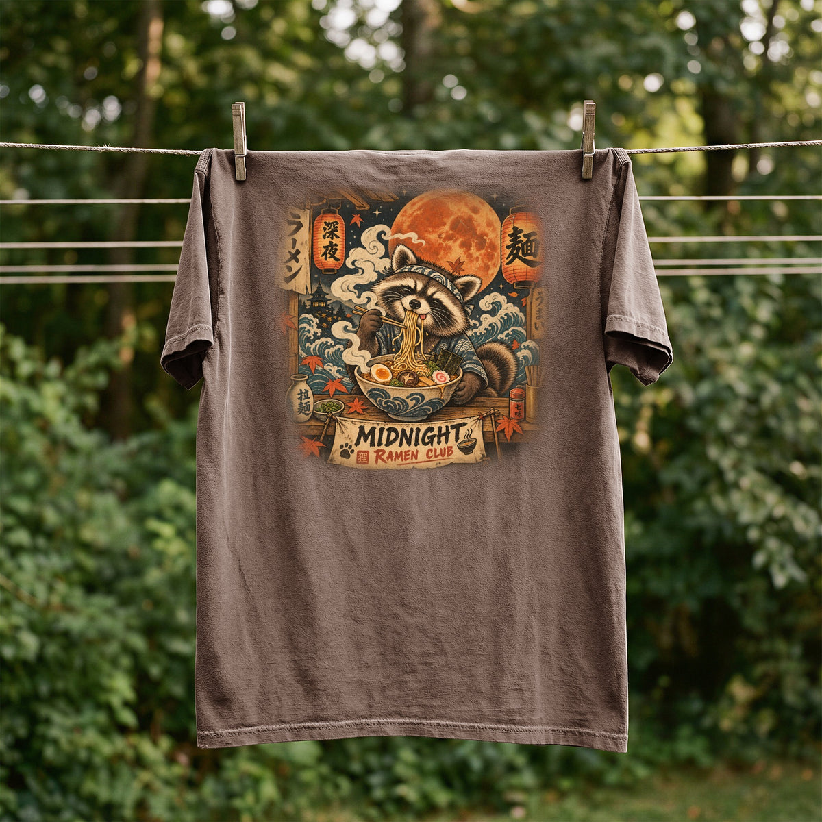 Brown t-shirt with graphic of raccoon eating ramen under a full moon.