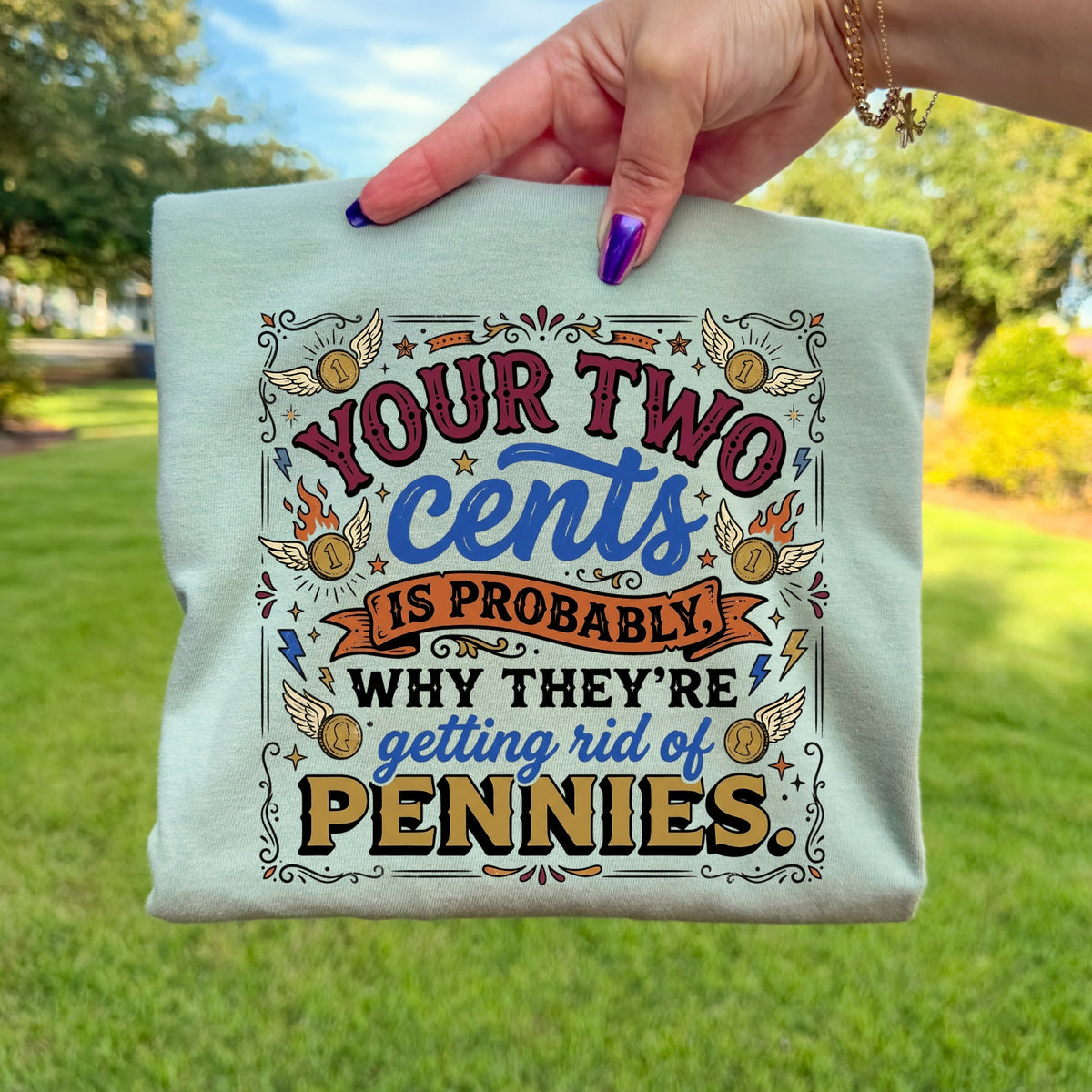A folded light green t-shirt held outdoors, featuring a retro colorful graphic with text reading Your Two Cents Is Probably Why They’re Getting Rid of Pennies.