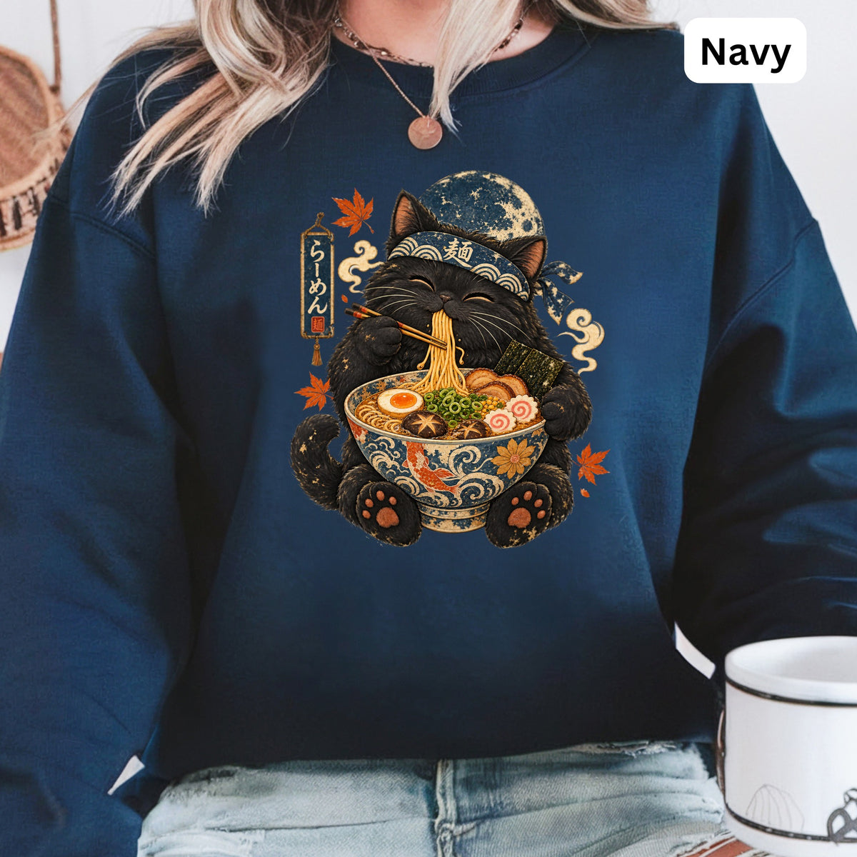 Black cat wearing a bandana eats ramen noodles from a bowl on a navy sweatshirt.