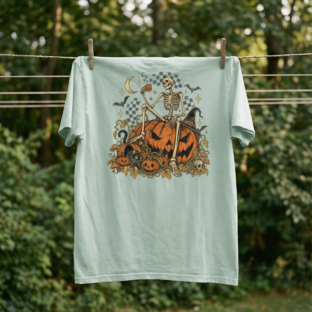 Skeleton with coffee cup on pumpkins, with moon, bats, and skull graphic on a t-shirt.