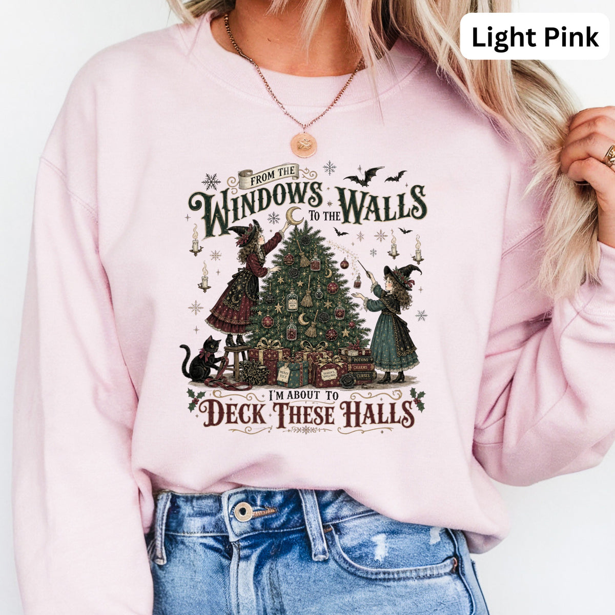 Light pink sweatshirt with witches decorating a Christmas tree and "Deck These Halls" text.