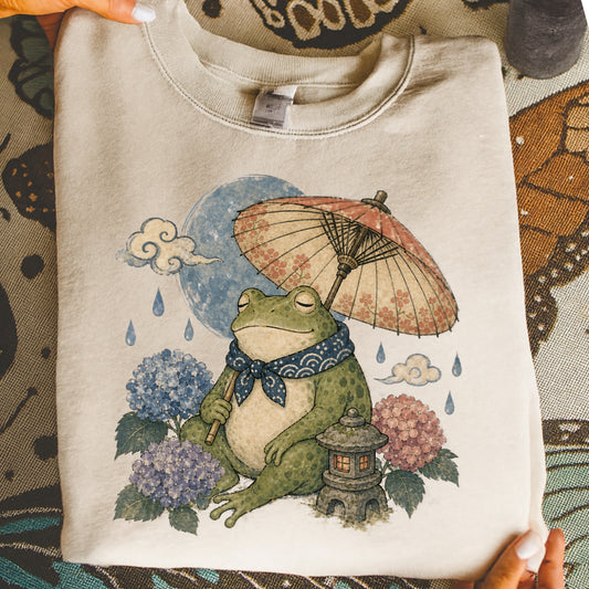 Frog holding umbrella with hydrangeas and a stone lantern on a light-colored sweatshirt.