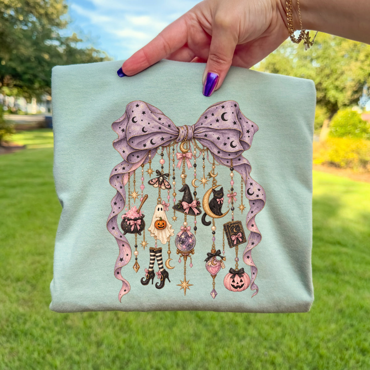 Teal shirt with a large purple bow adorned with moons and stars, featuring hanging Halloween-themed charms.