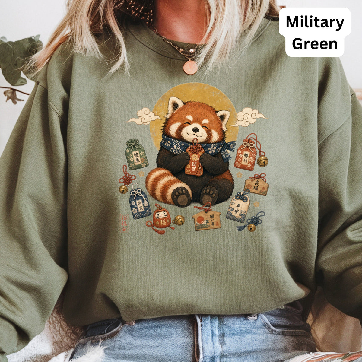 Red panda wearing scarf holding charm surrounded by Japanese talismans on green sweatshirt.