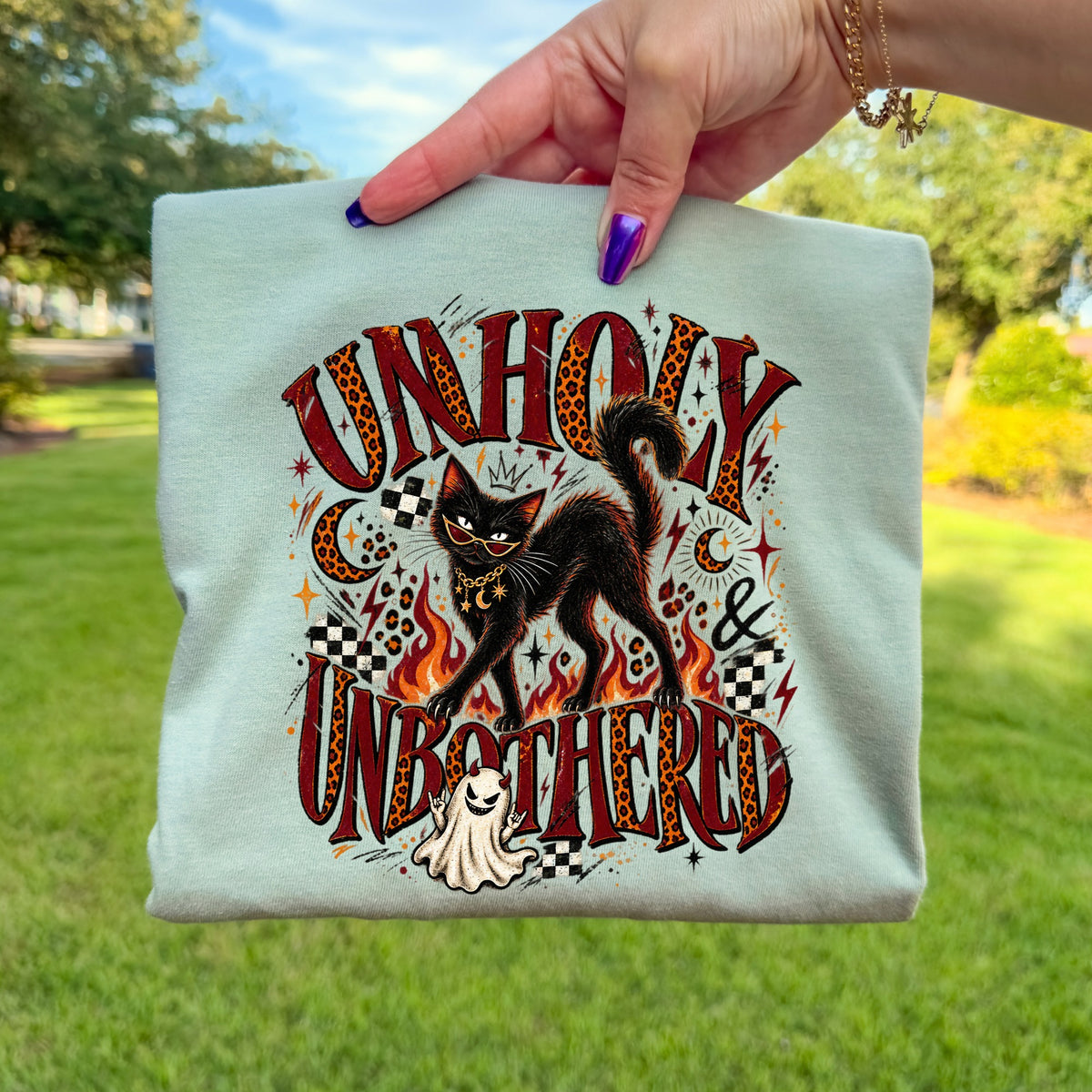 Light blue t-shirt with "Unholy & Unbothered" text, black cat, ghost, and flames graphic.