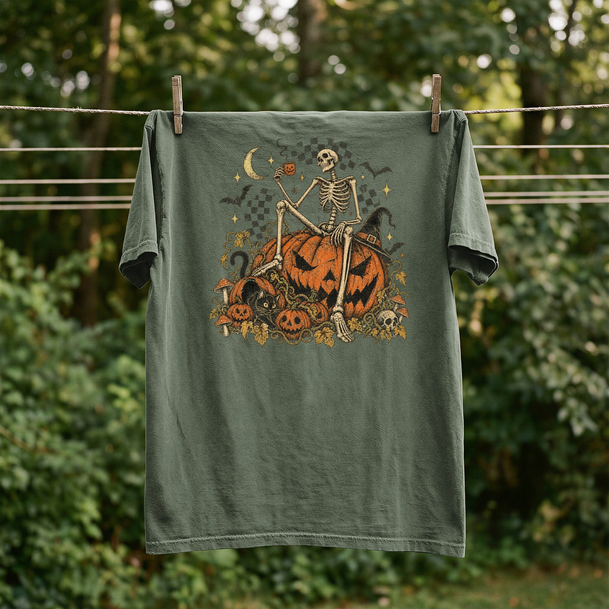 Skeleton sitting on pumpkins with moon, bats, and mushrooms on a green t-shirt.
