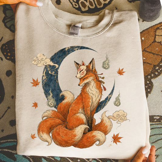 Cream sweatshirt featuring a nine-tailed fox with a mask sitting before a moon.